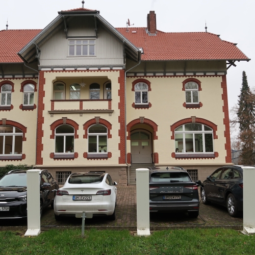 älteres, weißes Gebäude mit roten Rändern an den Fenstern und davor parkenden Autos